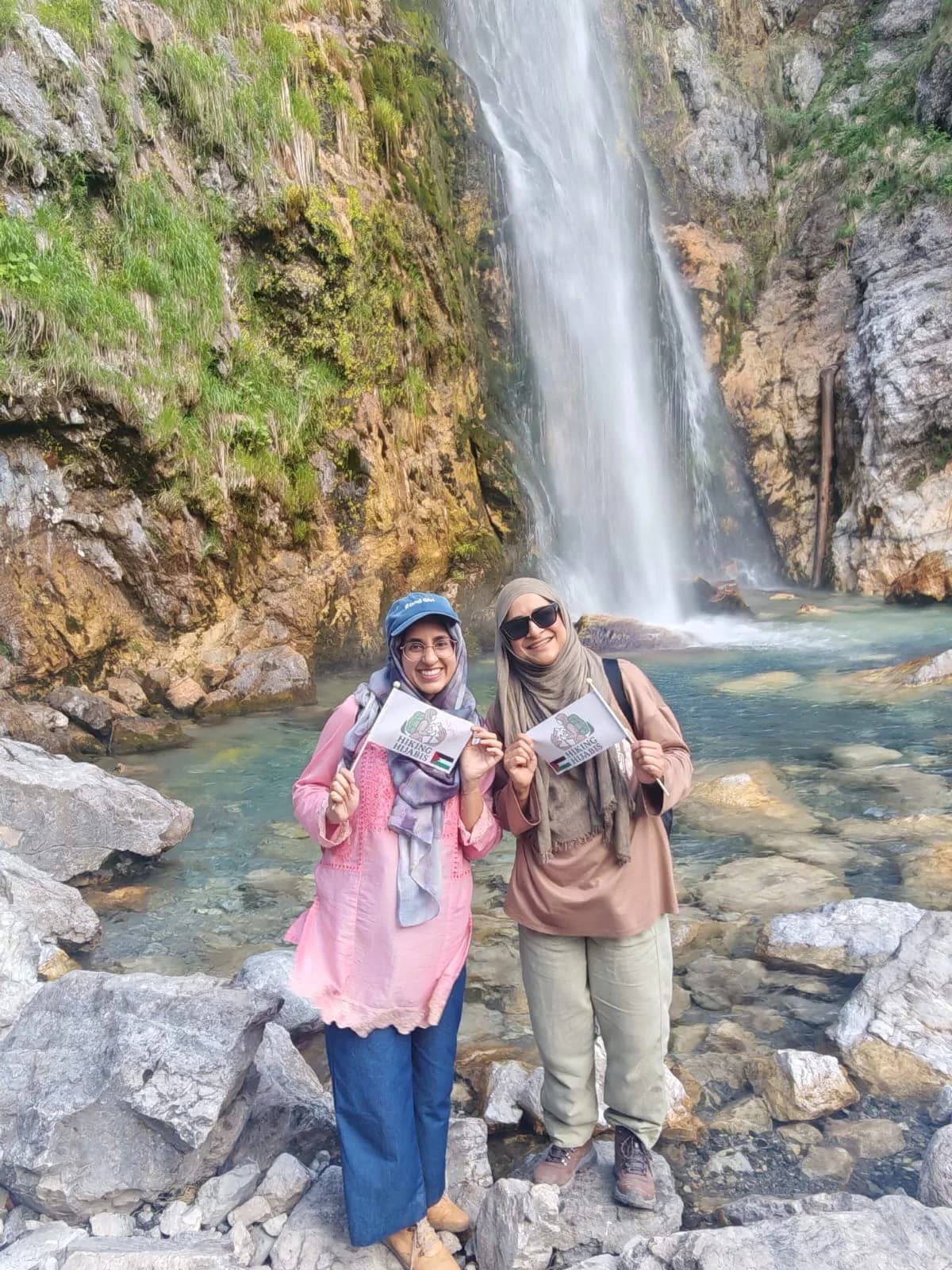 Two sisters smiling beside a waterfall and its clear pool